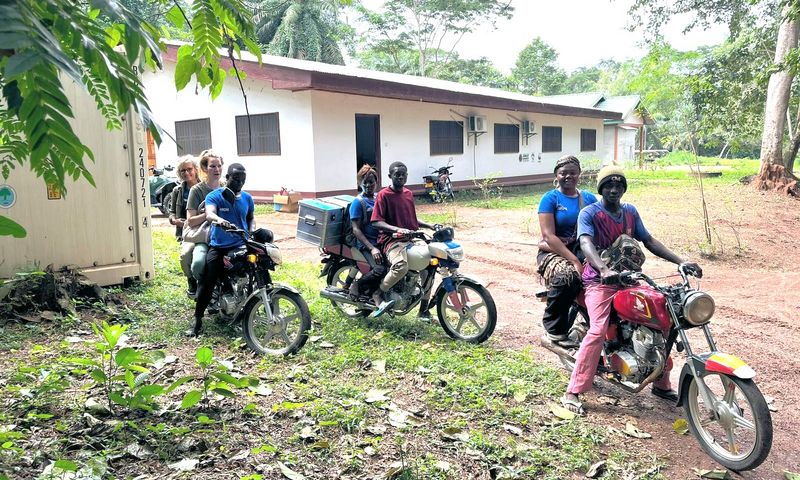 Gruppe von Menschen auf Motorrädern und Motorrollern vor einem weißen Gebäude in tropischer Umgebung.