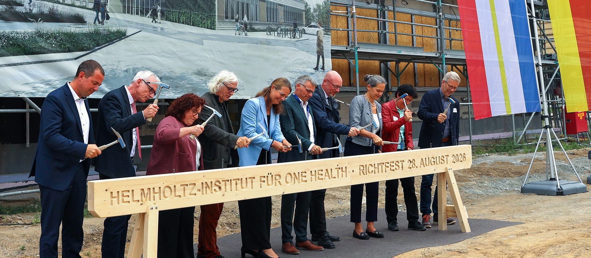 Neun Personen schlagen bei einem Richtfest auf einer Baustelle mit Hämmern symbolisch auf einen Holzbalken.