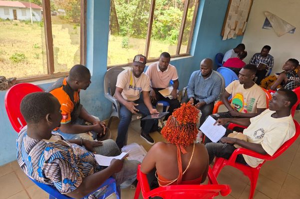 Gruppe von Menschen sitzen in Stuhlkreis, eine Person erklärt etwas an Tablet