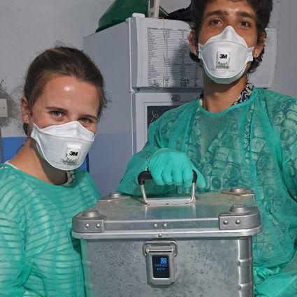 Two people in lab coats and masks are holding a silver box